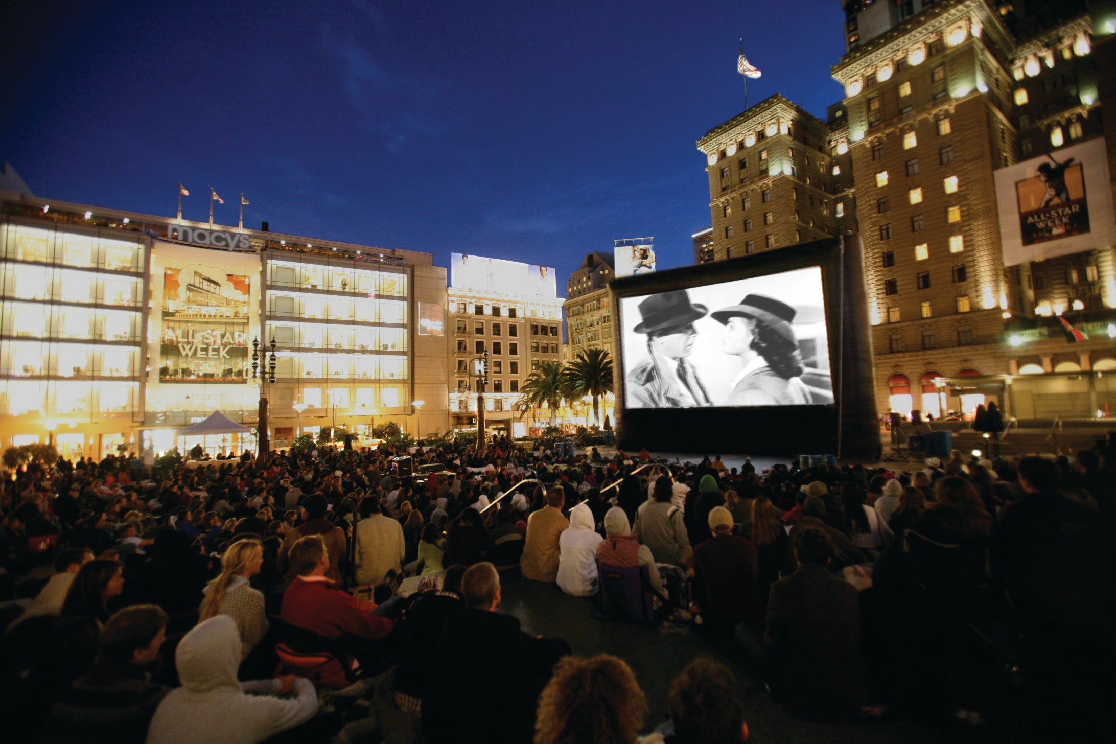 A crowd watching an Open Air Cinema Elite screen at night in a city square