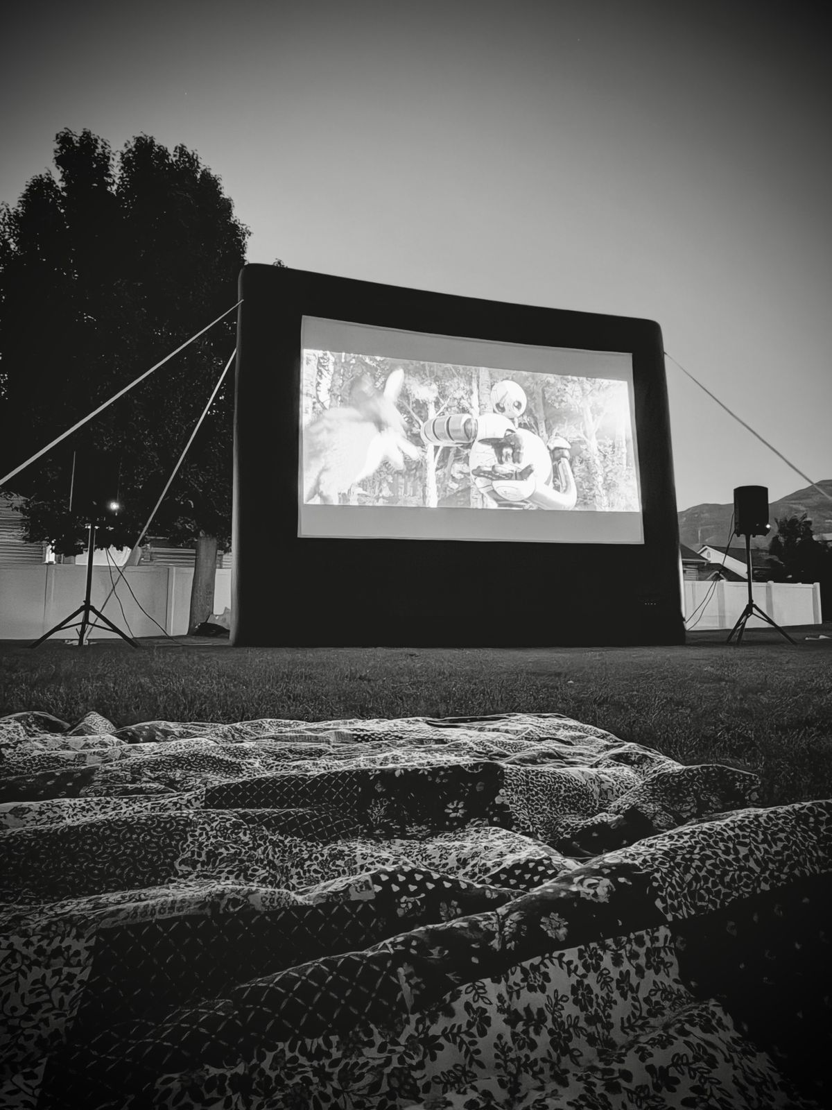Open Air Cinema Pro 12' inflatable screen showing a movie at dusk in a backyard, with speakers on stands and a quilt on the grass